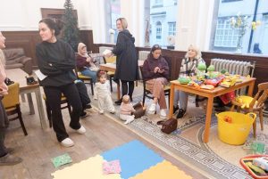 Women chatting casually in a room where toys and a child can also be seen.