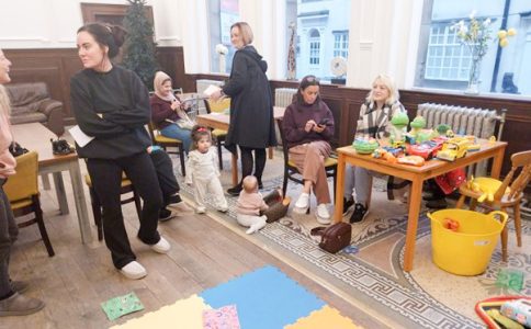 Women chatting casually in a room where toys and a child can also be seen.